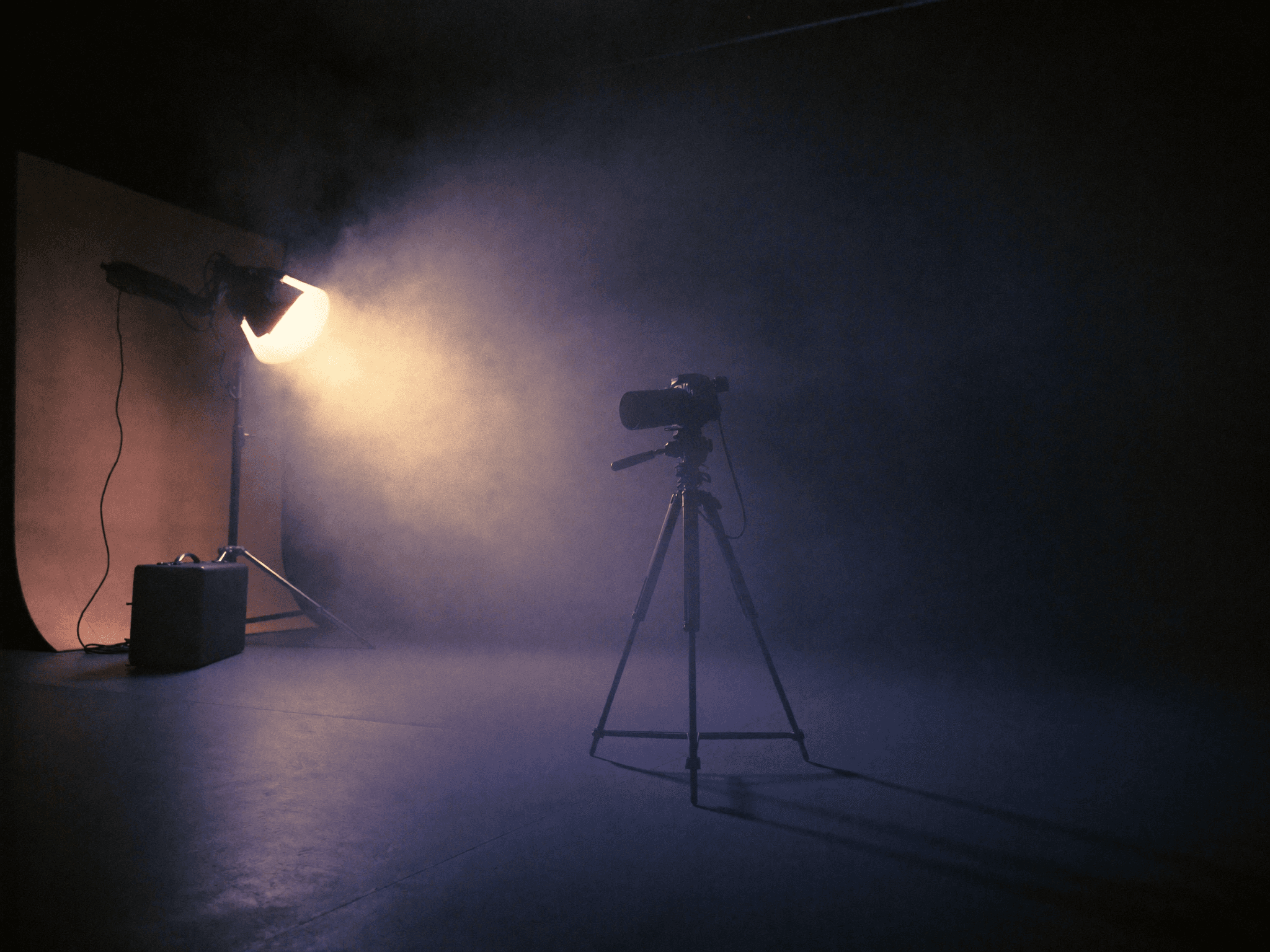A camera on a tripod beside a studio light in a smoke filled studio.